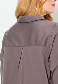 Camicia grigia con colletto, caratterizzata da un design con schiena plissettata. Tessuto liscio e leggero, che mette in mostra un taglio contemporaneo.