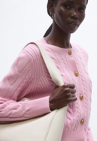 Rózsaszín kötött kardigán kábelmintával, arany rózsa alakú gombokkal és bordázott mandzsettával; egy krémszínű táskával kiegészítve.