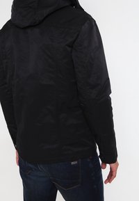 Sort hættejakke med en glat overflade, der har lynlås foran og elastiske manchetter. Sammen med blå denim jeans.