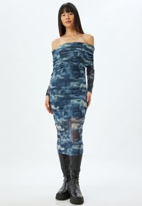 Vestido ajustado con hombros al descubierto en un patrón abstracto azul y verde, hecho de material elástico con detalles fruncidos, combinado con botas negras hasta la rodilla.