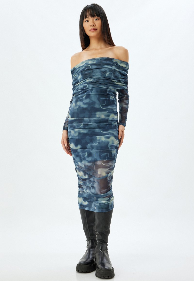 Vestido ajustado con hombros al descubierto en un patrón abstracto azul y verde, hecho de material elástico con detalles fruncidos, combinado con botas negras hasta la rodilla.