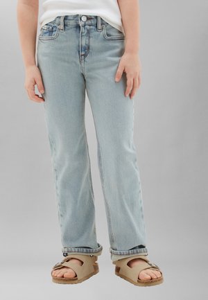 Enfant portant un jean bleu clair et des sandales beiges à bout ouvert, debout devant un fond gris uni.