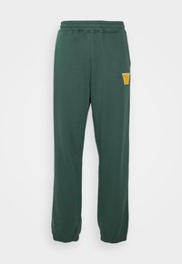 Gröna sweatpants i mjukt tyg, med en elastisk midja och muddars, samt en gul broderad 'W'-logotyp på vänster sida.