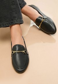 Loafers negri din piele, cu detalii dintr-un lanț aurit, purtați cu blugi închiși pe un fundal neutru. Pune accent pe încălțămintea elegantă și ținuta casual.