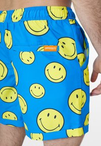Shorts de bain bleus avec un motif répétitif de visages souriants jaunes, taille élastique et étiquette orange "OPPOSUITS" sur la poche arrière.