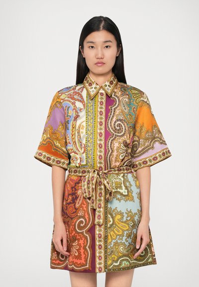 Femme portant une robe chemise à motif paisley coloré, à manches courtes et avec une ceinture à la taille, se tenant devant un fond blanc uni.