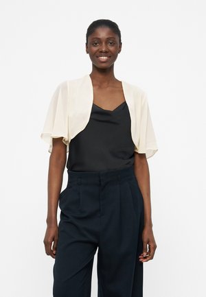 Χαμογελαστή γυναίκα που φοράει ένα κρεμ μπολερό με κοντά μανίκια πάνω από μαύρο μπλουζάκι και ψηλόμεσα μπλε σκούρα πλισέ παντελόνια, μπροστά σε λευκό φόντο.