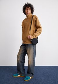 Brown zip-up fleece, loose blue denim jeans with distressed details, and teal sneakers with white accents. Black circular crossbody bag.