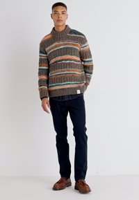 Suéter de punto con cuello alto en gris apagado, con rayas en naranja, azul verdoso y crema. Combinado con pantalones azul oscuro y zapatos marrones.