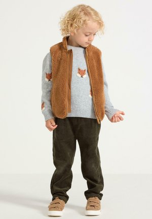 Bruine fleece bodywarmer over een grijze trui met vosontwerpen, gecombineerd met groene corduroypantalon en bruine sneakers met klittenbandsluitingen.