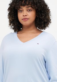 Lyseblå v-hals genser laget av mykt stoff, med lange ermer og en liten logo på brystet. Glatt tekstur med søm detaljer.