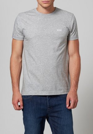 Homme portant un T-shirt gris clair ajusté à manches courtes avec le logo "BOSS" sur la poitrine gauche et un jean bleu foncé, debout devant un fond uni.