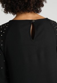 Haut noir avec un design de dos en trou de serrure, doté d'une fermeture à pression et de manches ornées de petites accents métalliques. Texture de tissu lisse.