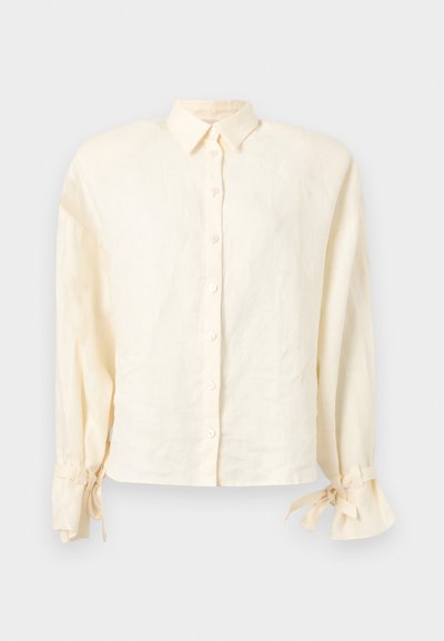 Κρεμ πουκάμισο με μακριά μανίκια, κουμπιά, αιχμηλό γιακά και δεμένες φιόγκους στους μανσέτες πάνω σε φωτεινό φόντο.