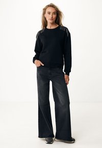 Schwarzer Sweatshirt mit Fransen-Details an den Schultern, kombiniert mit lockeren schwarzen Jeans und grauen Sneakers. Glatter Stoff, lässige Passform.