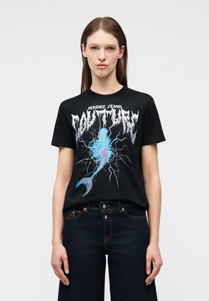 Γυναίκα που φοράει μαύρο T-shirt με μπλε και ροζ γραφικό γοργόνας και κείμενο "Versace Jeans Couture", συνδυασμένο με σκούρο μπλε τζιν.