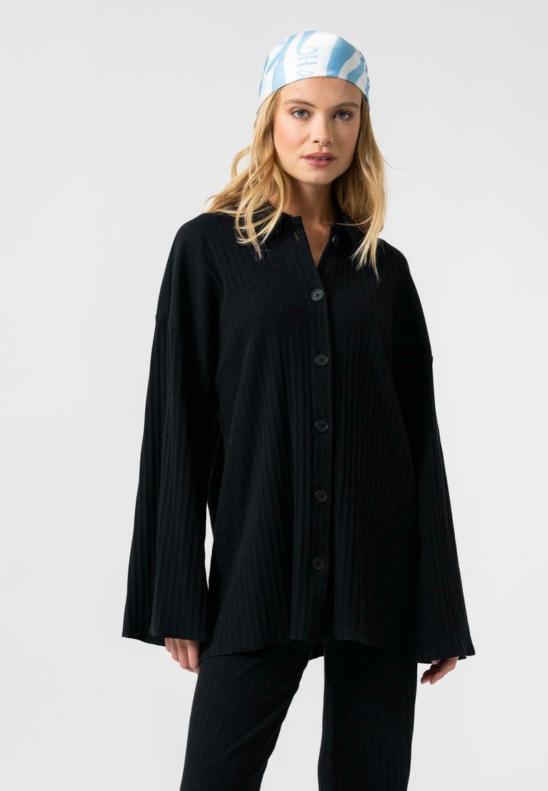 Zwarte geribbelde knoopshirt met lange mouwen, voorzien van een puntige kraag en zijsplitten, gecombineerd met bijpassende zwarte broek.