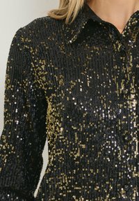 Camicia nera e dorata in paillettes con colletto classico, caratterizzata da una superficie testurizzata e un motivo splendente di paillettes.