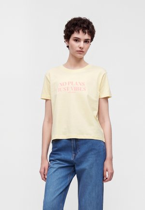 Jeune personne aux cheveux courts et bouclés portant un t-shirt jaune pâle avec le texte « NO PLANS JUST VIBES » et un jean bleu, debout devant un fond blanc.