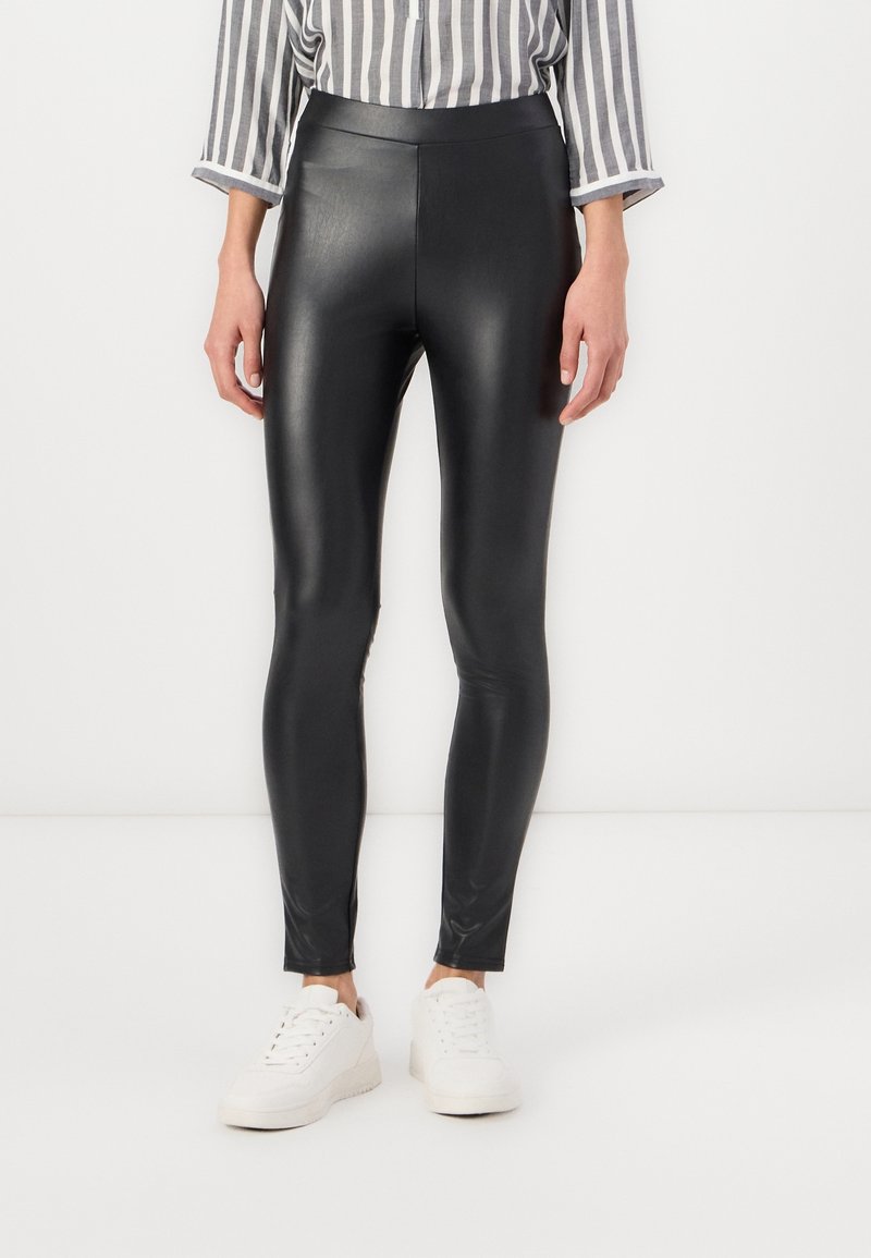 Personne portant des leggings noirs en simili cuir, des baskets blanches et une chemise rayée verticale gris et blanc, debout devant un fond blanc.