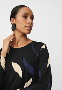 Zwarte top met lange mouwen met abstracte beige en blauwe vormen. Model draagt grote zwarte geometrische oorbellen, die een gladde textuur en modern ontwerp tonen.