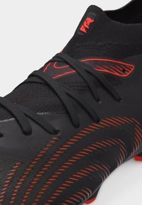 Μαύρο ποδοσφαιρικό παπούτσι με κόκκινες λεπτομέρειες, υφασμένο επάνω μέρος, μαύρα κορδόνια και κόκκινα ραφές σε λευκό φόντο.