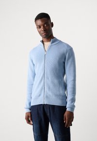 Light blue zip-up sweater with ribbed texture, high collar, and side pockets, paired with dark denim pants. Close-fitting design.