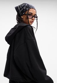 Crna oversized hoodie s mekanom teksturom, s kapuljačom na vezanje. Nosena s uzorkovanom crnom bandanom i elegantnim pravokutnim sunčanim naočalama.