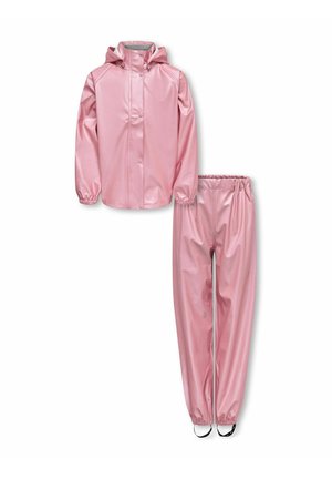 Jeune fille portant une veste à capuche rose brillante et un pantalon assorti, debout les bras croisés et bottes noires devant un fond uni.