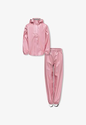 Jeune fille portant une veste à capuche rose brillante et un pantalon assorti, debout les bras croisés et bottes noires devant un fond uni.