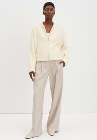 Crème faux fur zip-up jasje met brede mouwen, gecombineerd met lichtbeige plooibroek en lichtgekleurde schoenen. Soepele, zachte textuur.