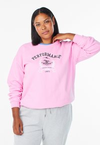 Svetlo roza majica z dolgimi rokavi, z napisom "PERFORMANCE" in grafiko. Kombinirana s sivimi trenirkami. Mehka, teksturirana tkanina.