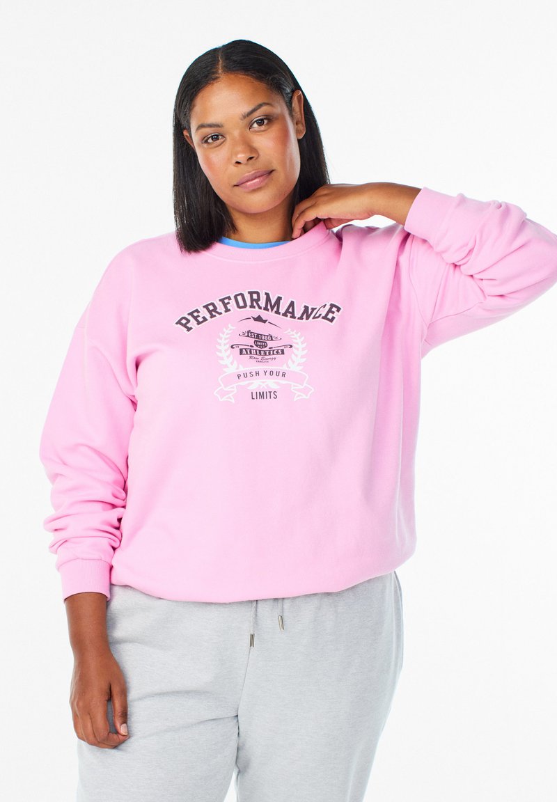 Svetlo roza majica z dolgimi rokavi, z napisom "PERFORMANCE" in grafiko. Kombinirana s sivimi trenirkami. Mehka, teksturirana tkanina.