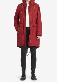 Rode parka met een capuchon, voorzien van een rits en knoopsluiting, twee voorkbakken en een trekkoord in de taille. Draagt over een wit sweater en zwarte jeans.