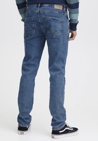 Uske slim-fit plave traperice izrađene od trapera, s pet džepova, prepoznatljivim detaljima šivanja i standardnim pojasom. Nosene s crnim tenisicama.