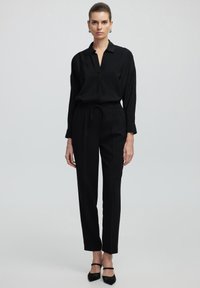 Blusa nera a maniche lunghe con patta nascosta, abbinata a pantaloni neri a vita alta con coulisse e gamba affusolata.