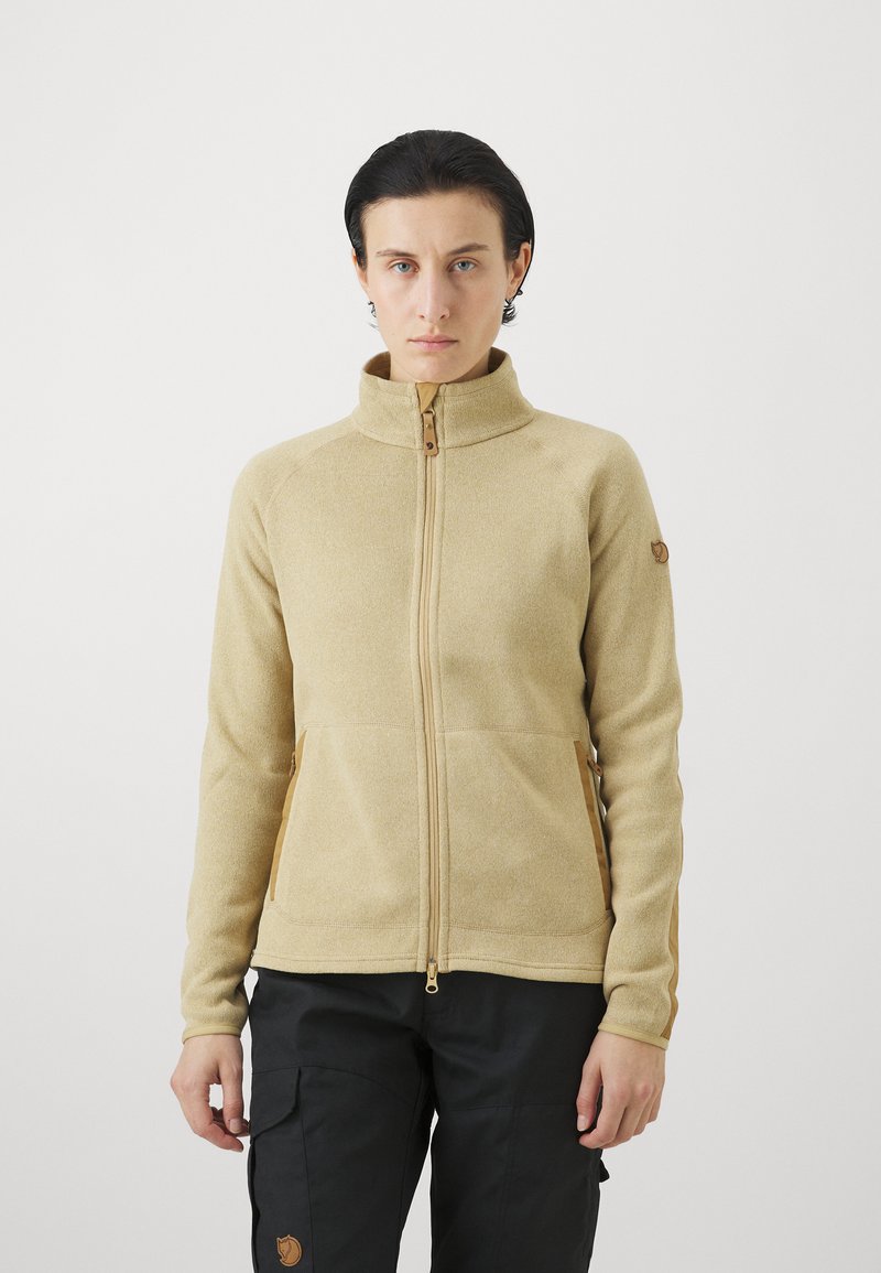 Giacca in pile beige con colletto alto, chiusura con zip e due tasche laterali. Presenta un tessuto texturizzato e una toppa con logo sulla manica.
