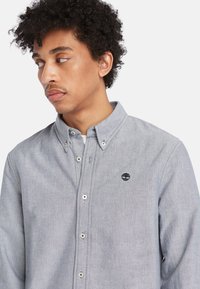 Camisa de botón gris confeccionada en un tejido texturizado, con cuello clásico, botones blancos y un pequeño logo oscuro en el pecho.