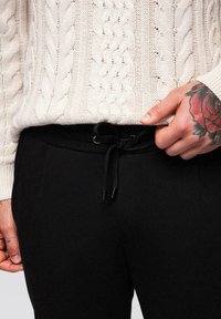 Gros plan sur une personne portant un pull en tricot crème et un pantalon noir à cordon de serrage, avec un tatouage de rose rouge sur la main droite.