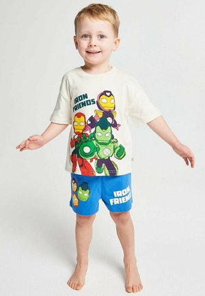 Lächelnder Junge steht barfuß und trägt ein weißes T-Shirt sowie blaue Shorts mit bunten Cartoon-Figuren der Iron Friends.