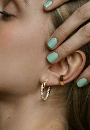 Guldøreringe med klare ædelstene, der har et snoet design. En guld cuff-ørering bæres over hoops, med grøn neglelak synlig.