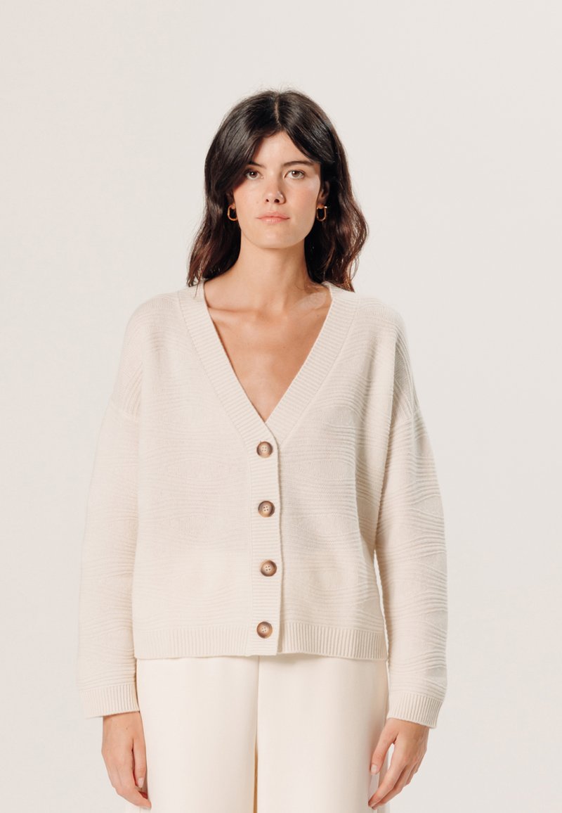 Ecrufarbener Strickcardigan mit tiefem V-Ausschnitt, fünf Holzknöpfen und gerippten Bündchen. Verfügt über ein strukturiertes Muster über den Stoff.