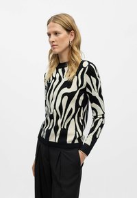 Pull en tricot noir et crème à rayures façon zèbre, avec un col rond, des manches longues et des poignets côtelés, associé à un pantalon noir taille haute.
