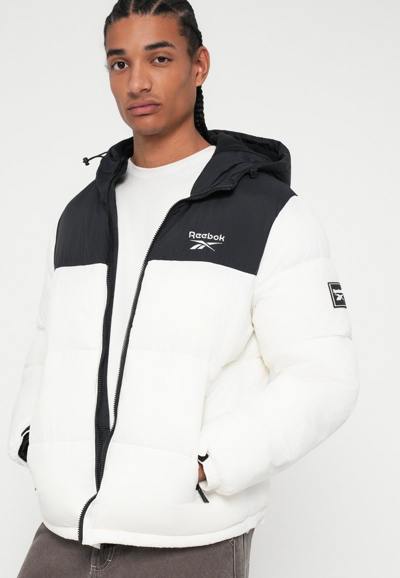Biała i czarna kurtka puchowa z kapturem, z pikowanym wzorem, przednim zamkiem błyskawicznym oraz logo Reebok na klatce piersiowej i rękawie.