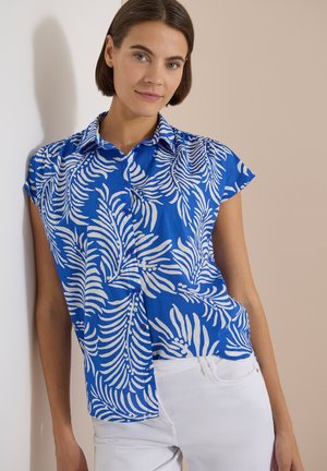 Femme aux cheveux bruns courts portant une chemise bleue avec des motifs de feuilles blanches et un pantalon blanc, s'appuyant contre un mur beige.