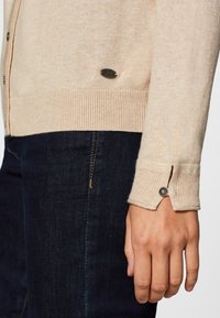 Beige knitted cardigan with ribbed cuffs, featuring a button detail and a small branded patch near the hem. Paired with dark denim jeans.