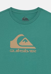 Tyrkysové bavlnené tričko s veľkým béžovým logom Quiksilver, ktoré má motívy hôr a vln. Klasický dizajn s okrúhlym výstrihom a krátkymi rukávmi.