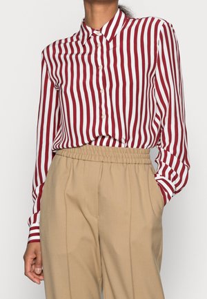 Mujer vistiendo una camisa abotonada con rayas verticales rojas y blancas, metida dentro de unos pantalones beige de cintura alta con una pretina elástica.