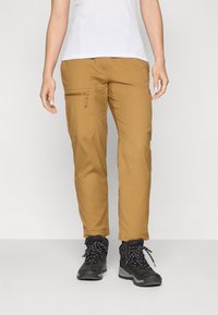 Κρεμ παντελόνια πεζοπορίας με φερμουάρ στη μηρή, φτιαγμένα από ελαφρύ ύφασμα, σε συνδυασμό με μαύρες μπότες πεζοπορίας με λευκές κορδόνες.