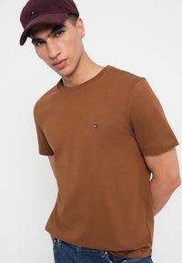 Καφέ βαμβακερό T-shirt με στρογγυλή λαιμόκοψη και κοντά μανίκια, που διαθέτει μια μικρή λογότυπη ετικέτα. Φορεμένο με μια μπορντό καπέλο από κορδόνι.